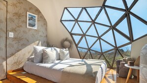 A bedroom with a large geometric window, a bed with white bedding, and a view of the outdoors. - Bosque Geométrico (Tepoztlán)