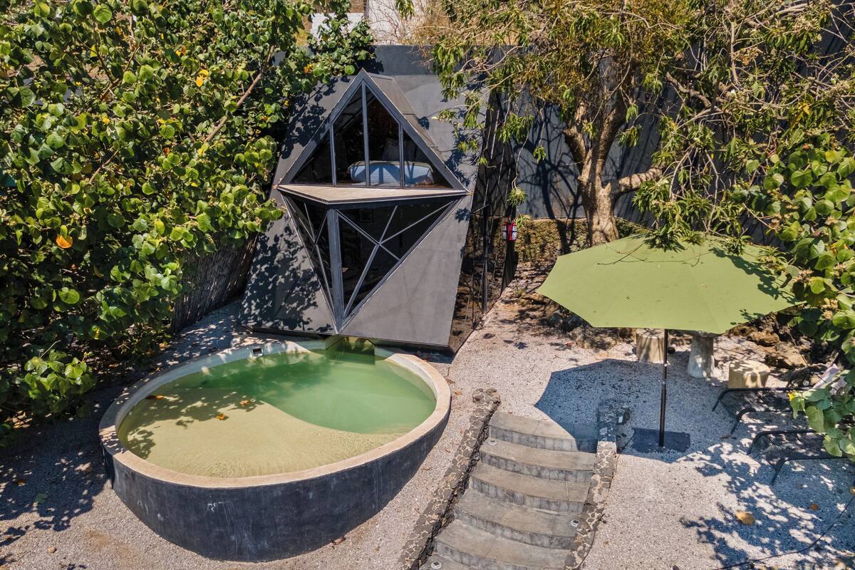 Una piscina circular con borde de piedra, una estructura geométrica y una escalera que lleva a una zona sombreada.