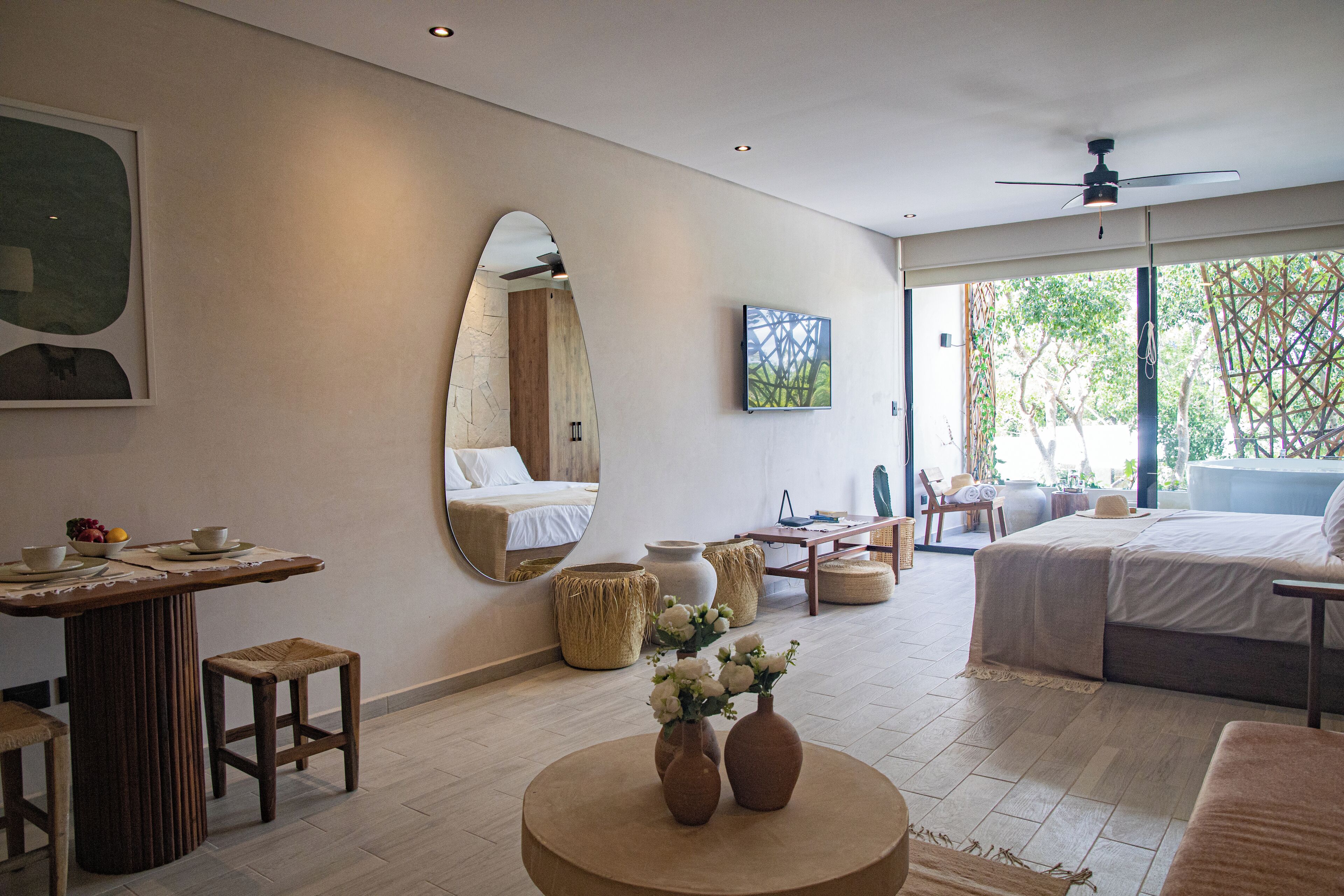 A modern hotel room with a large bed, a dining area with a table and chairs, a round coffee table with vases, and a large mirror on the wall.