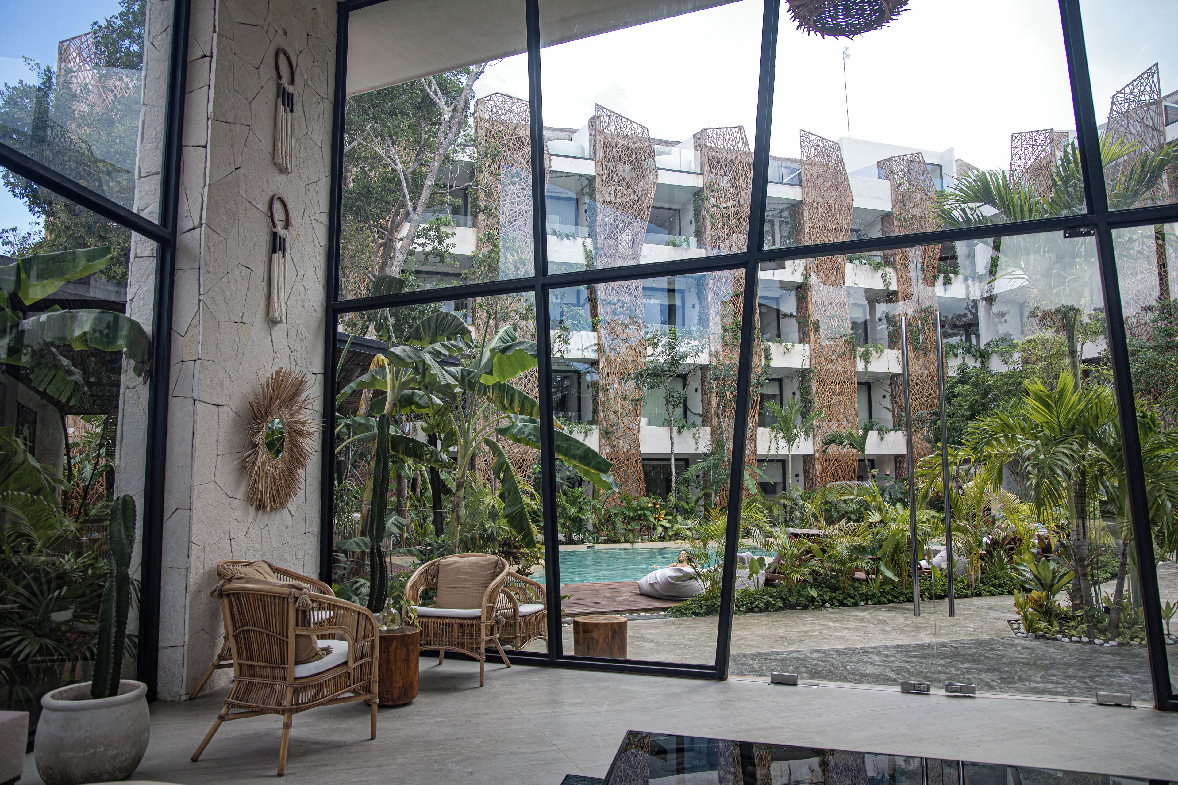 Un espacio interior moderno con ventanas amplias, mobiliario de mimbre y vistas a una piscina y follaje.