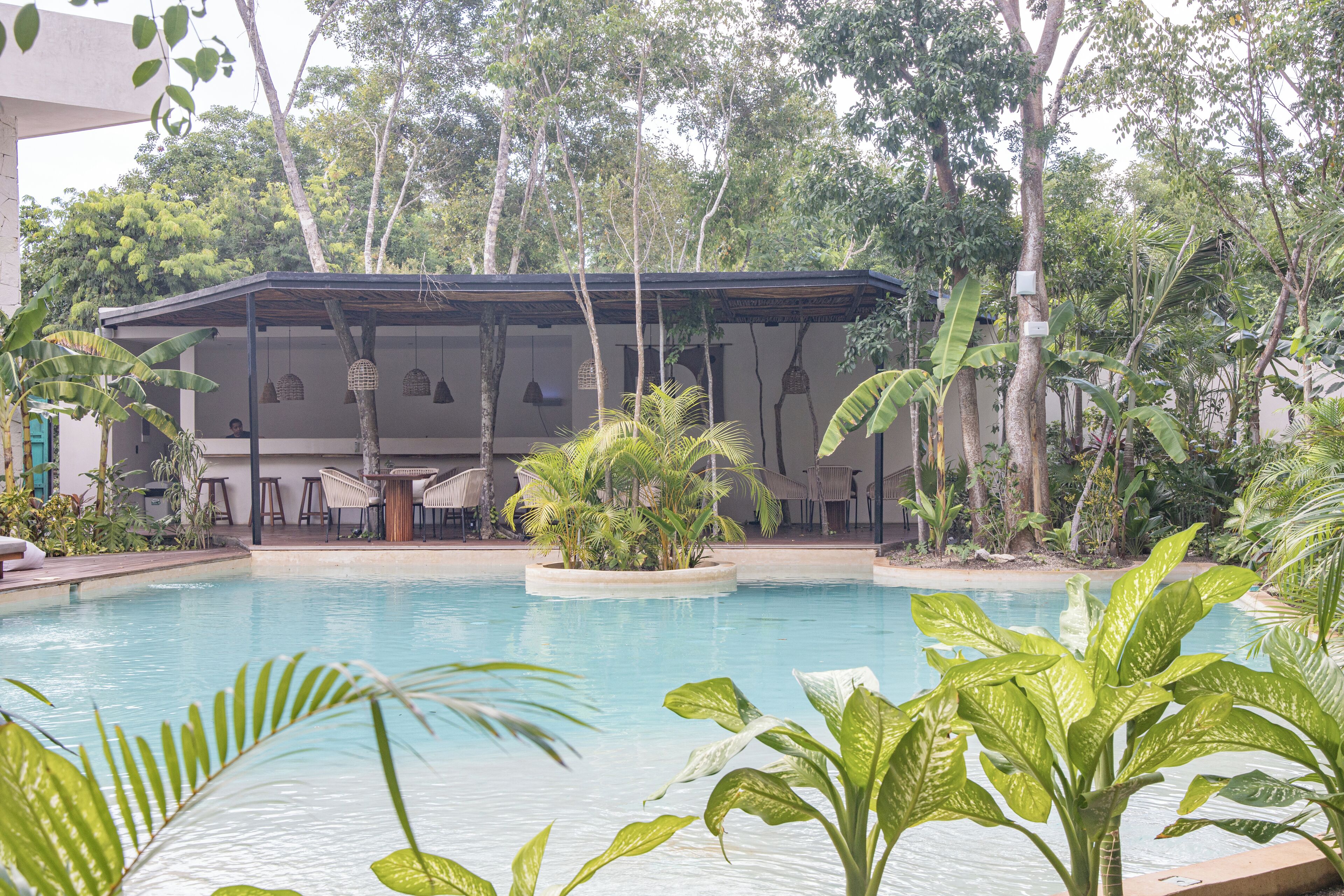 A pool area with a covered seating area, surrounded by lush greenery and trees.