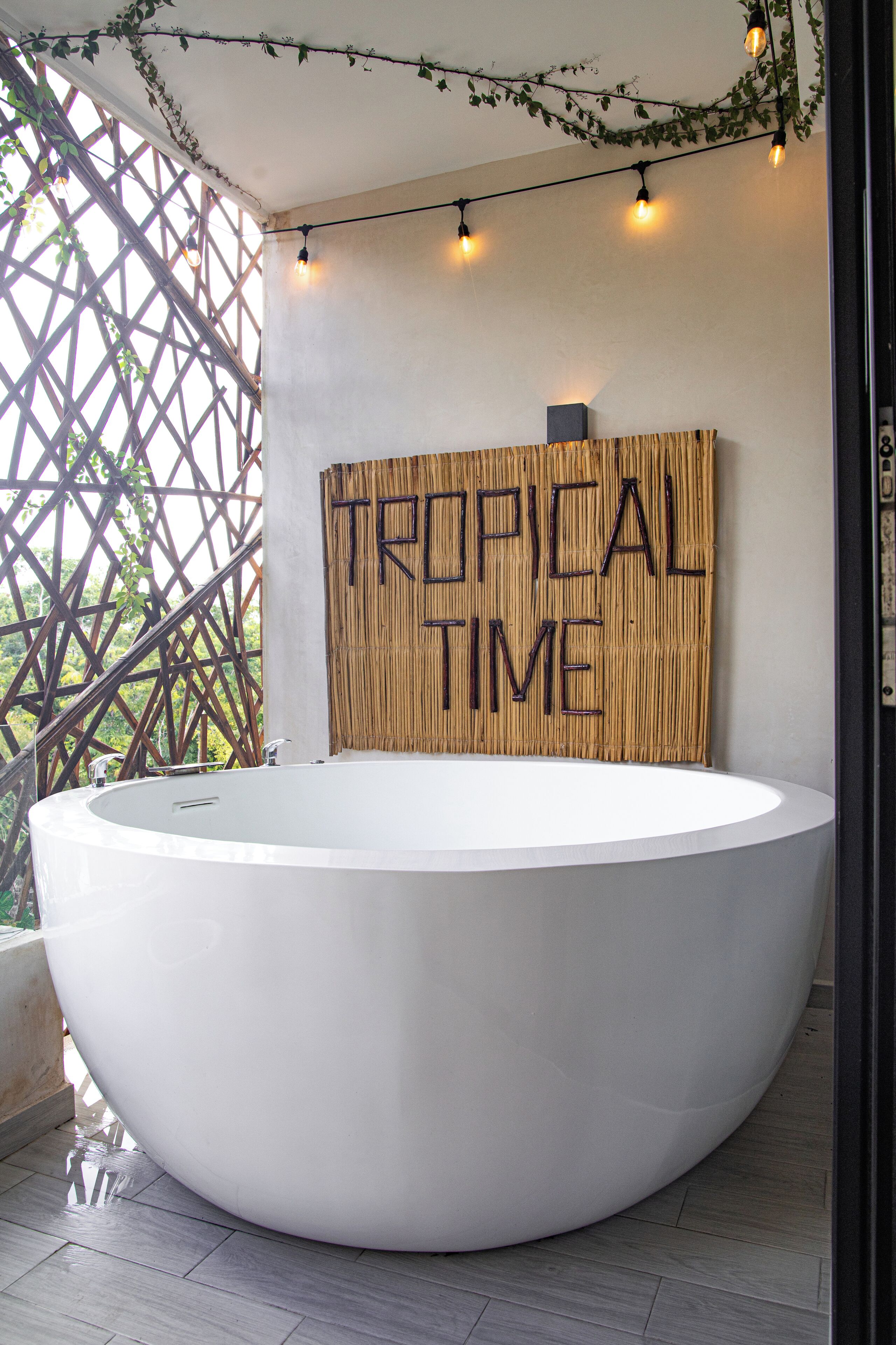 Un baño moderno con una bañera blanca grande, un muro de bambú con el texto 'TROPICAL TIME' y luces colgantes.