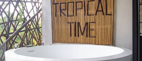 Un baño moderno con una bañera blanca grande, un muro de bambú con el texto 'TROPICAL TIME' y luces colgantes.