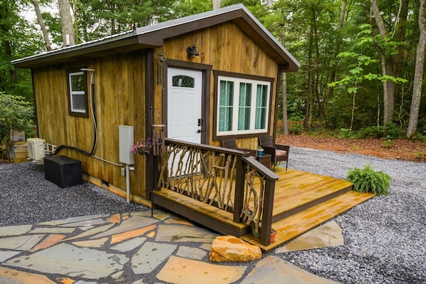 Une cabane en bois avec une entrée pavée de pierres, un petit porche doté d’une balustrade et une porte surmontée d’une fenêtre.