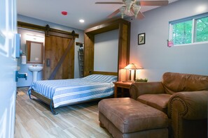 A bedroom with a bed, a wooden wardrobe, a bathroom with a sink and mirror, a brown leather chair, and a window with a view of trees.