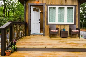 Una terraza de madera con una puerta blanca, dos sillas de mimbre y plantas en macetas.