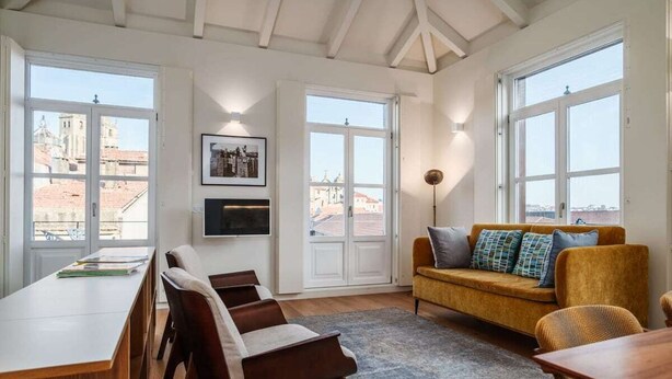 A spacious room with a long wooden desk, a comfortable sofa with cushions, and large windows offering a view of buildings. - T2 Flores (Porto)