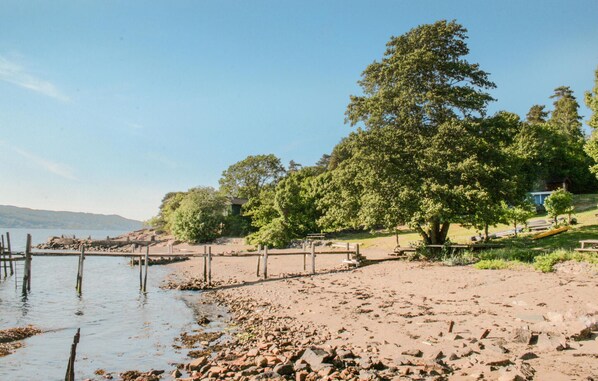 Een strand met een houten pier die uitsteekt in rustig water, omringd door bomen en een gazon met picknicktafels.