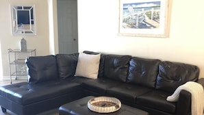 Una sala de estar con un sofá seccional de cuero negro, una mesa de centro redonda tejida, un tapete mullido, un cuadro en la pared y una pequeña mesita auxiliar con un objeto decorativo.