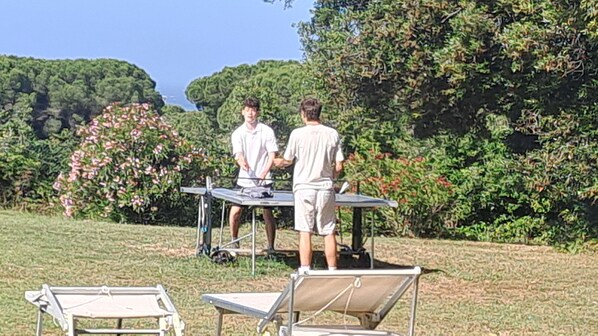 Dos personas están de pie junto a una mesa al aire libre, con una zona verde y árboles de fondo.