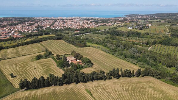An aerial view of a coastal town with terraced fields, a prominent red-roofed building, and a distant cityscape. - AGRITURISMO LE CERBONCHE (Rosignano Marittimo)