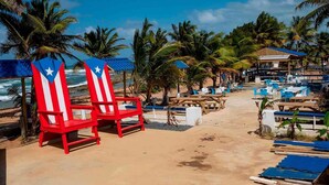 A beachside resort with red and white striped chairs, tables, and umbrellas, surrounded by palm trees and a view of the ocean.