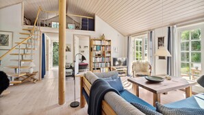 Een ruime loft-achtige kamer met een hoge plafond, een houten trap, een grote boekenkast, een comfortabele bank en een eettafel.