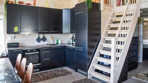 Une cuisine moderne avec des meubles de couleur sombre, un lave-vaisselle et une cuisinière encastrée. Un escalier en bois se trouve sur la droite, et il y a un coin repas avec une table et des chaises.