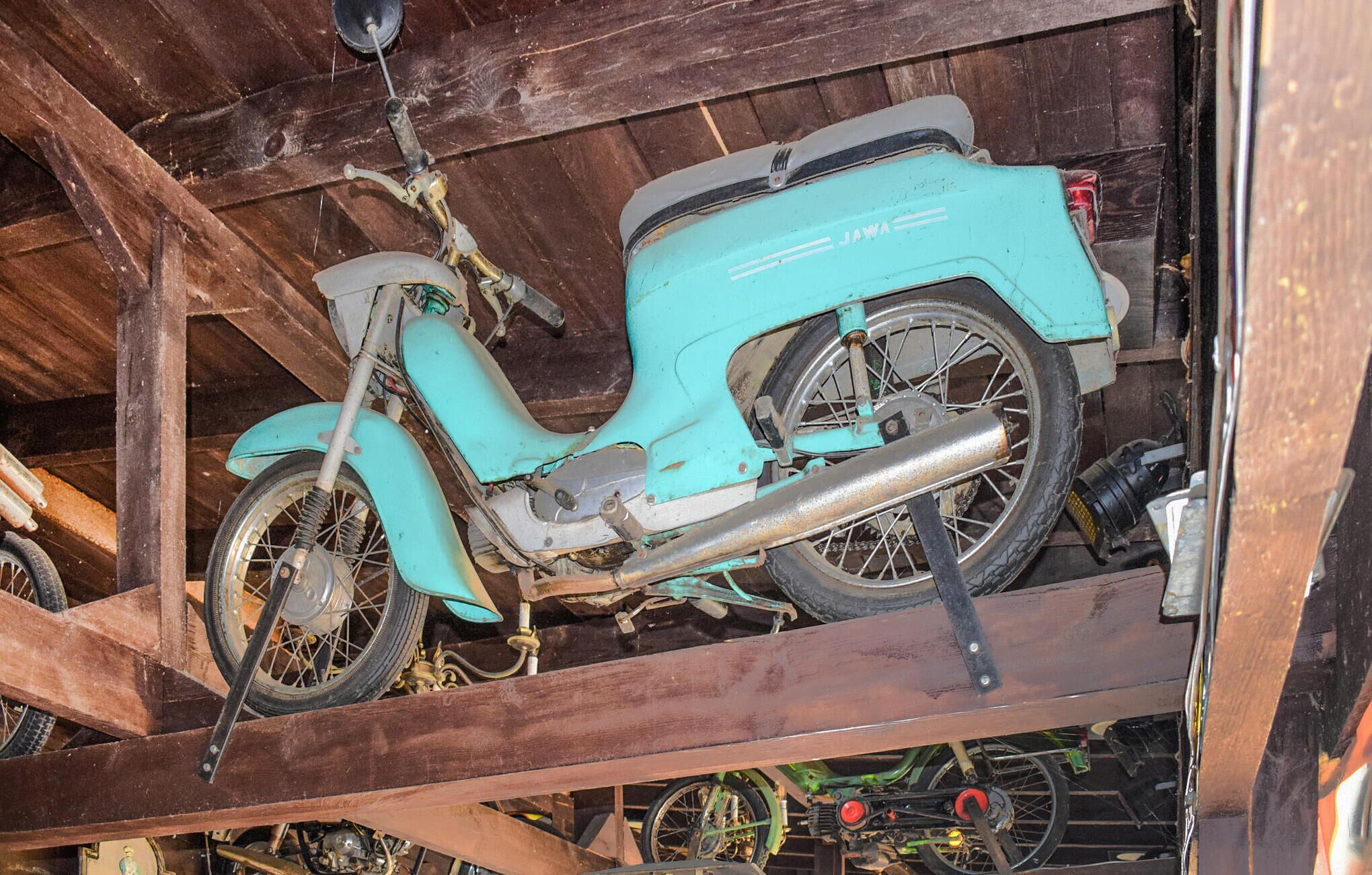 A vintage teal scooter with visible wear and rust, stored in a wooden structure with other bicycles.