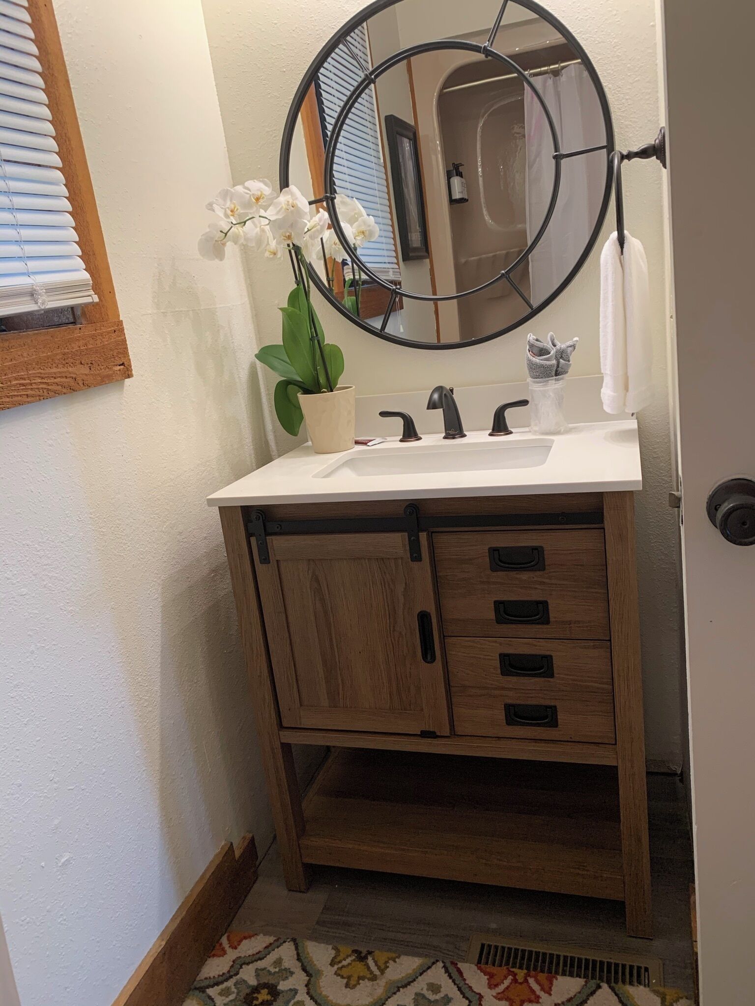 A bathroom with a wooden vanity, a round mirror, a white sink, and a potted orchid.