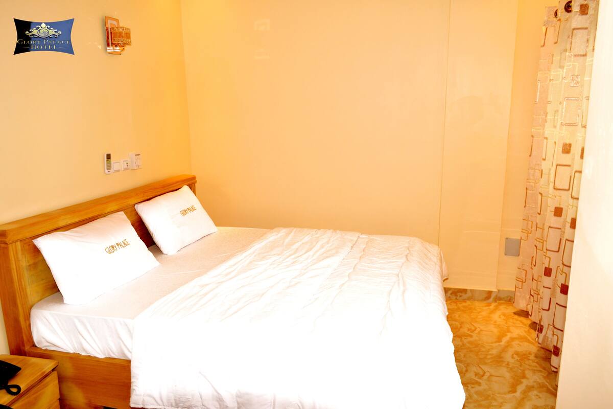A hotel room with a wooden bed, white bedding, a nightstand, and a wall-mounted light switch.