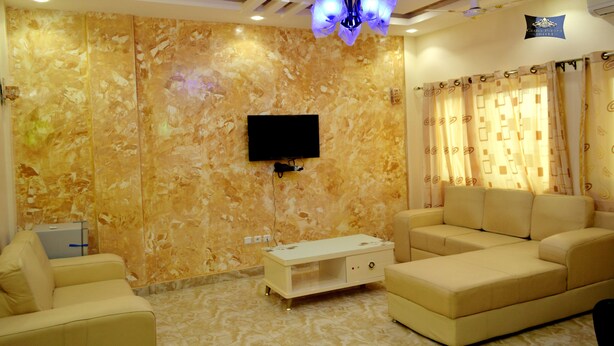 Ein Wohnzimmer mit Flachbildfernseher an einer Marmorwand, beigefarbenen Sofas, einer weißen Unterhaltungseinheit und gemusterten Vorhängen.