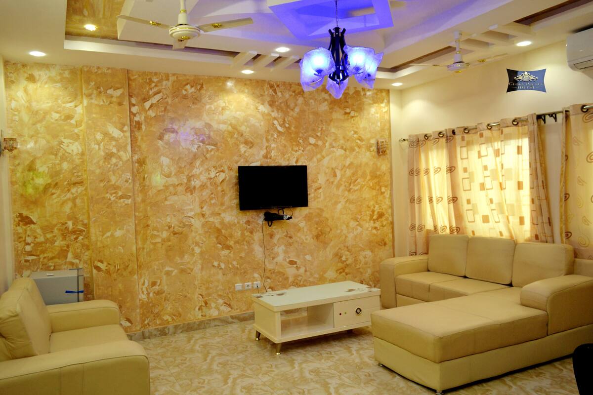 A living room with a flat-screen TV mounted on a marble wall, beige sofas, a white entertainment unit, and patterned curtains.