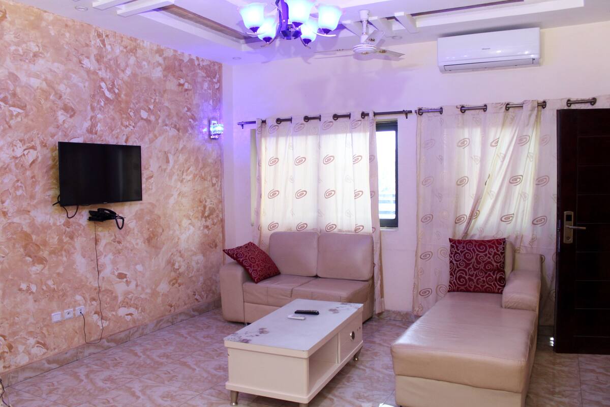 A living room with a sofa, a coffee table, a TV mounted on the wall, and a window with patterned curtains.