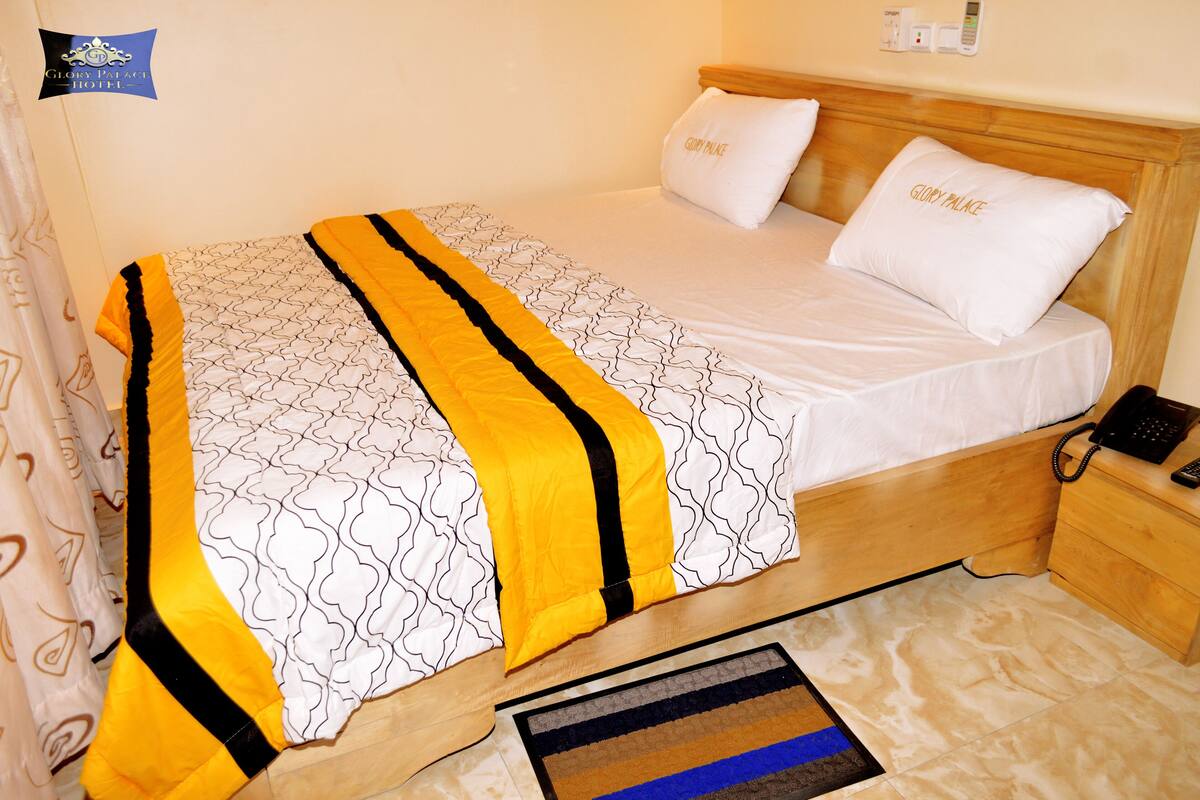 A neatly made bed with a patterned bedspread, two pillows with the hotel's name, a bedside phone, and a small wooden nightstand.