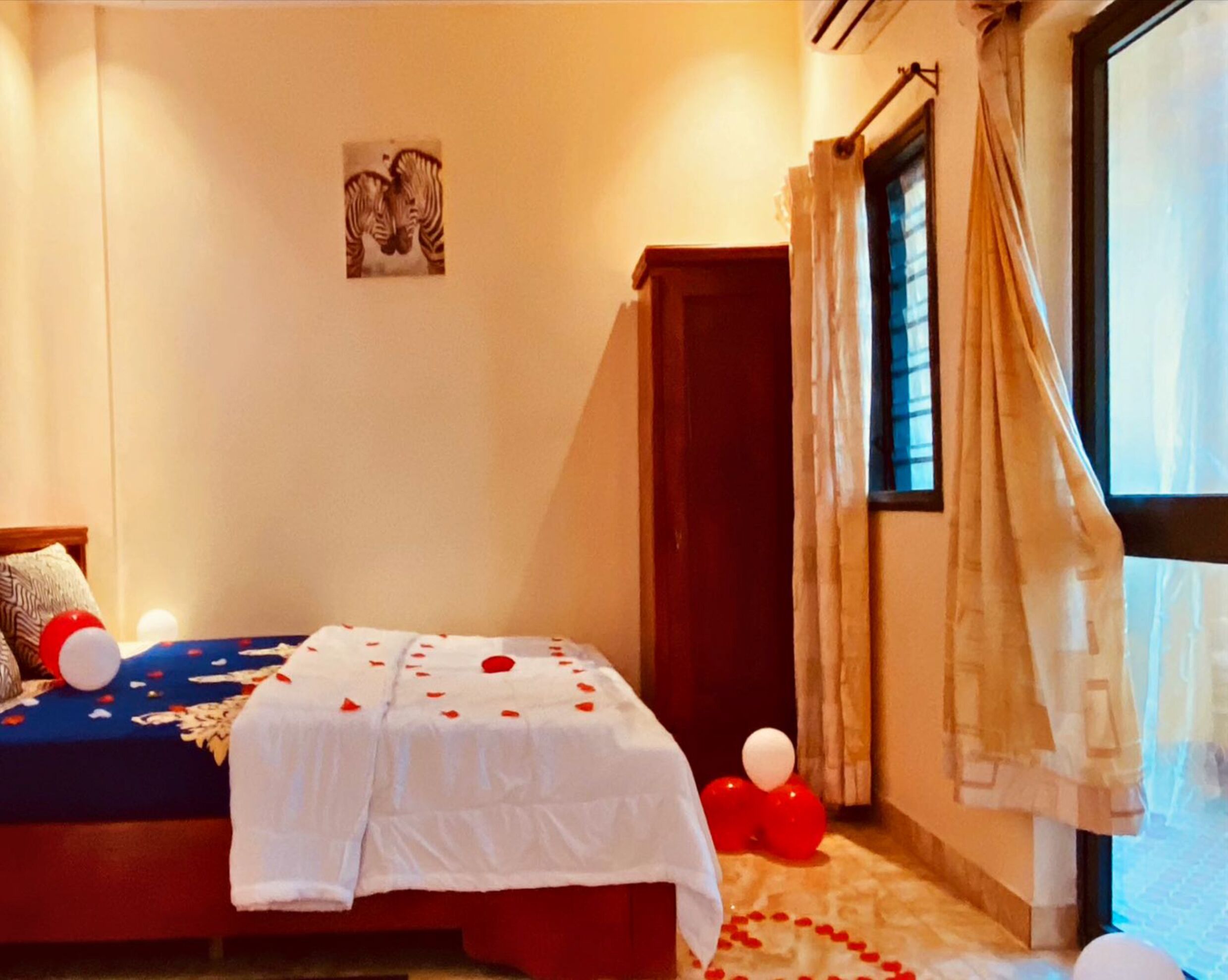 A bedroom with a bed, a wall-mounted picture, a window with curtains, and a small window with a view of the outside.