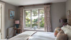 Ein Schlafzimmer mit einem großen Bett, floralen Vorhängen, Blick ins Grüne, einem Heizkörper und einem gerahmten Bild an der Wand.