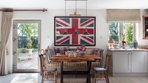 Ein Essbereich mit Holztisch, Stühlen und einer Union-Jack-Flagge darüber. Ein Fenster bietet Blick auf Grünfläche.
