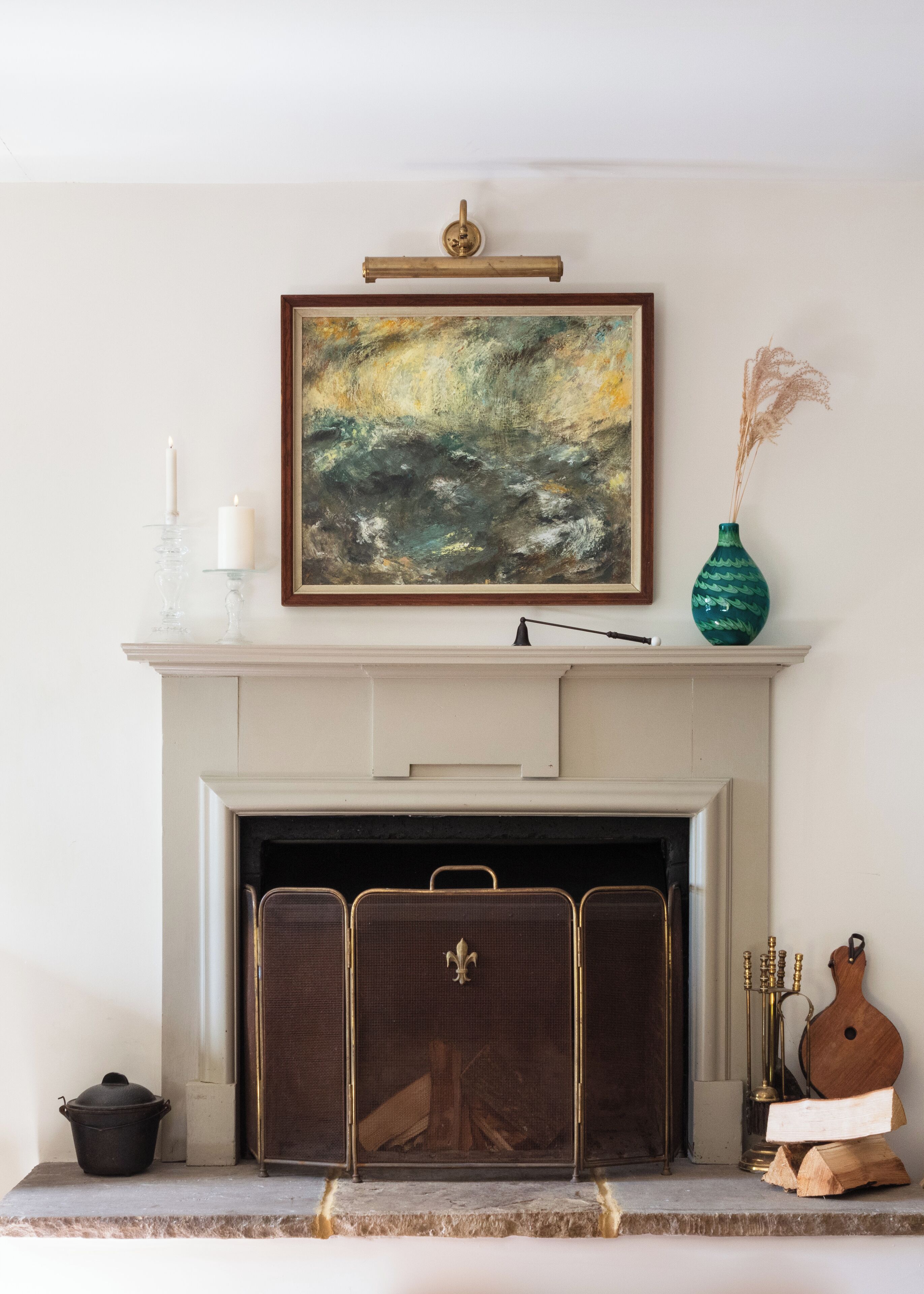 A fireplace with a framed abstract painting above it, a vase with dried plants, and a decorative stand with candles.