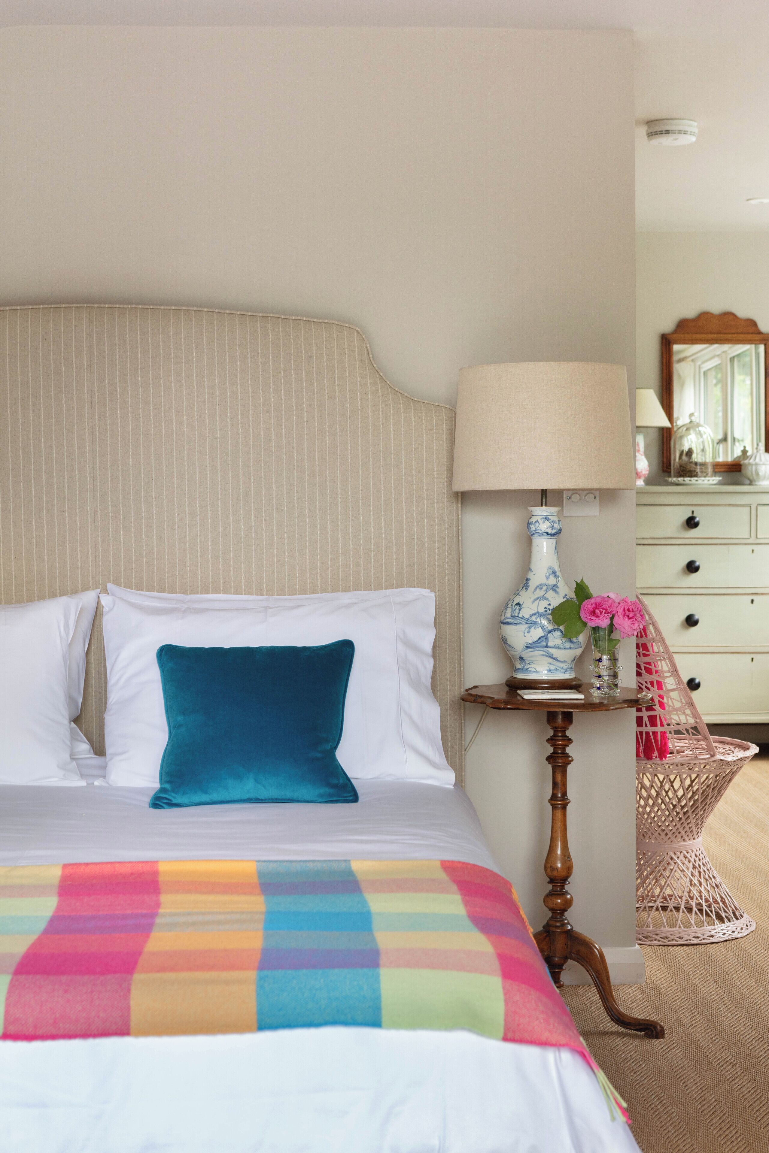 A neatly made bed with a colorful blanket, a teal pillow, and a bedside table with a lamp and vase of flowers.