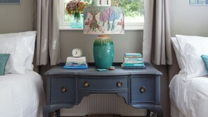 A bedroom with two beds, a dresser, a window with curtains, a vase with flowers, and a painting on the wall. - Nest Lodge, a stunning three bedroom lodge surrounded by nature (Pulborough)