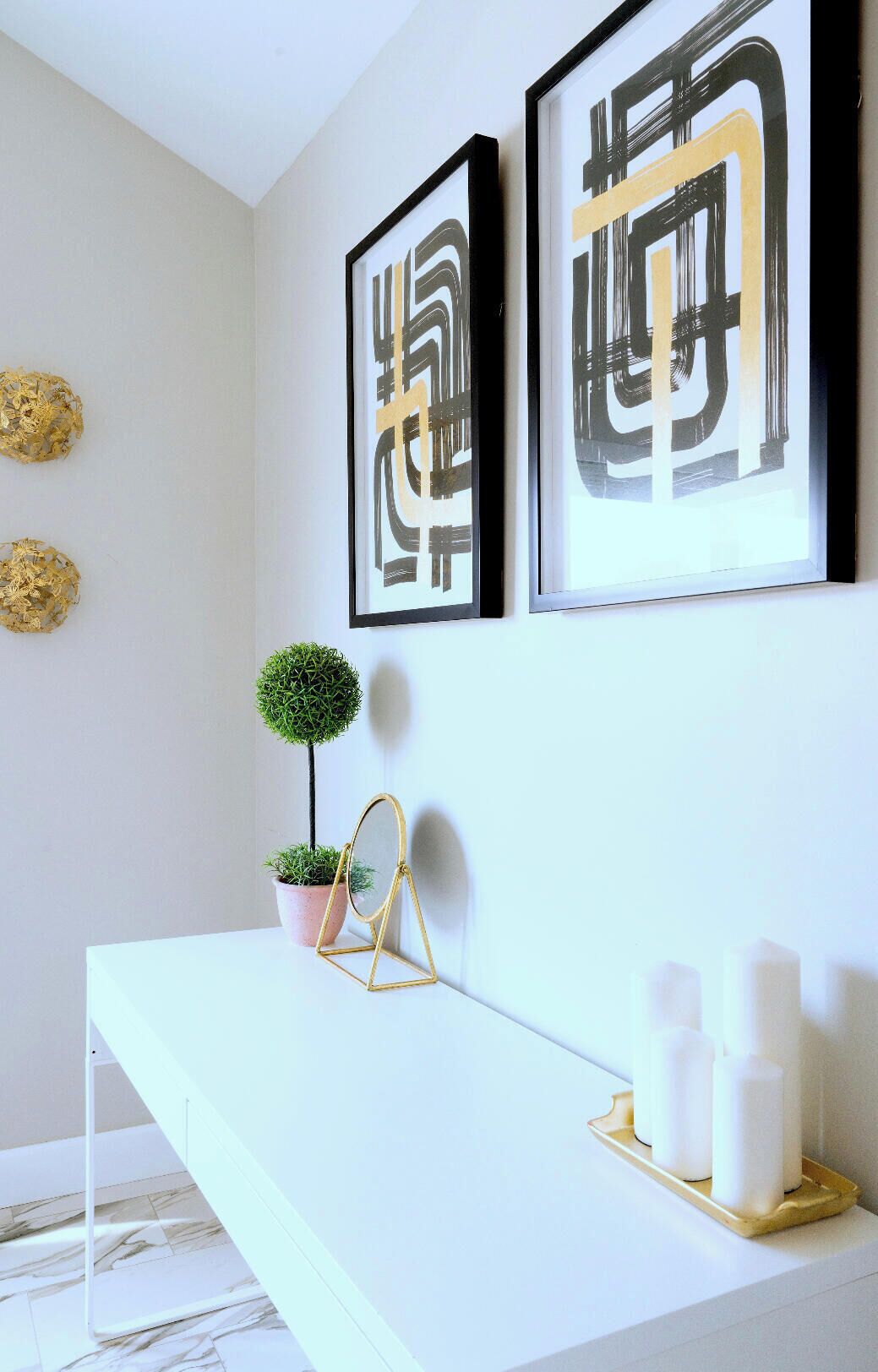 Een minimalistisch ingerichte kamer met een witte console tafel, twee kunstwerken met abstracte motieven, een kleine potplant en een gouden decoratie aan de muur.