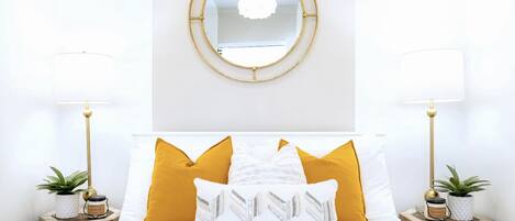 Un dormitorio ordenado con una cama centrada entre dos mesitas de noche, cada una con una lámpara y una planta. Un espejo redondo cuelga sobre la cama.