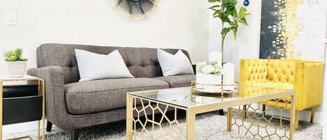 A living room with a grey sofa, a yellow armchair, a glass coffee table, and a decorative mirror on the wall.