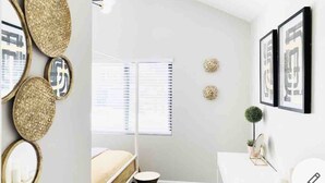 Un dormitorio moderno con cama, mesita de noche y una pequeña planta en maceta. Hay cuadros en la pared y una ventana con persianas.