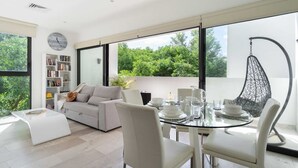A modern living room with a glass dining table, white furniture, a bookshelf, and a large window with a view of greenery. - Mexication Beach Bliss: Stylish Playa Getaway (Playa del Carmen)
