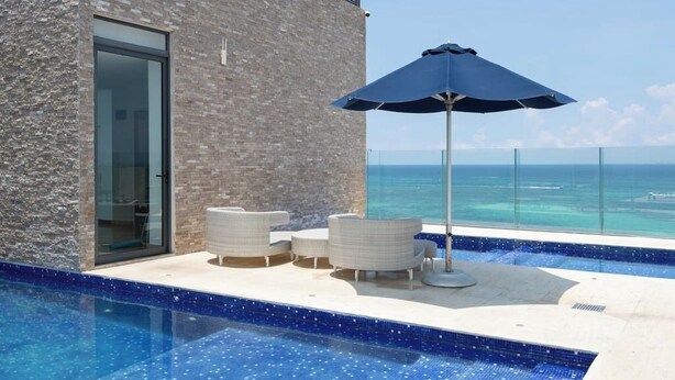 A modern outdoor pool area with a clear view of the ocean, a glass door entrance, and outdoor seating. - Mexication Beach Bliss: Stylish Playa Getaway (Playa del Carmen)
