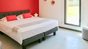 Ett modernt sovrum med en stor säng, röda accentväggar och utsikt över ett grönskande landskap genom ett stort fönster.