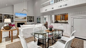 A spacious living room with a glass-top dining table, white wicker furniture, a fireplace, and a large TV.
