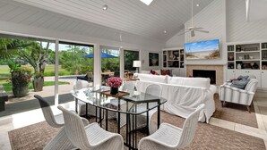 A spacious living area with a glass dining table, white wicker furniture, a fireplace, and a large TV.