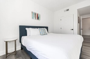 A neatly arranged bedroom with a large bed, a small side table, and a door leading to another room. - Beach Condo 436CB (Carlsbad)
