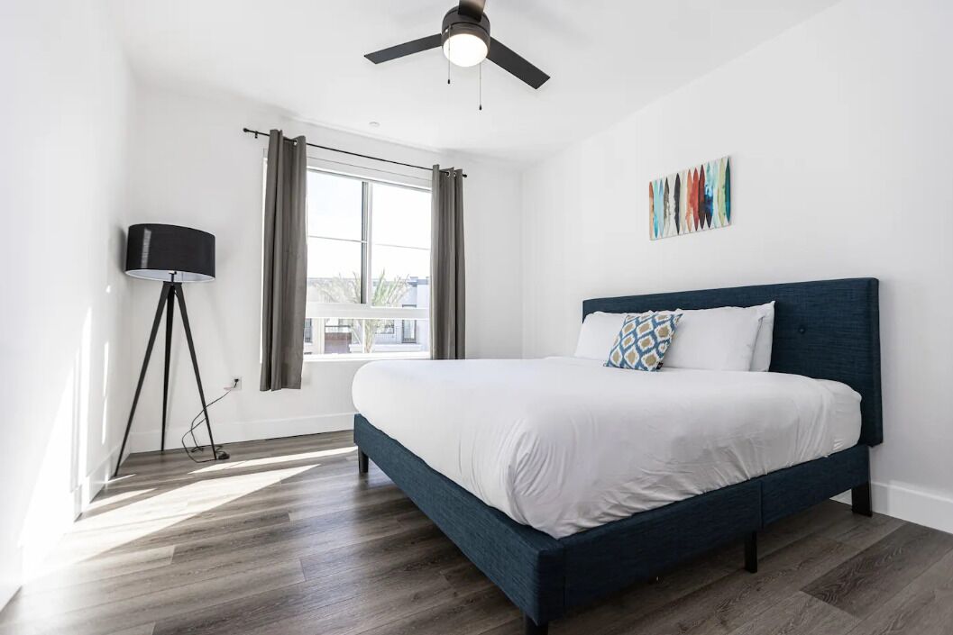 A bedroom with a bed, a standing lamp, a ceiling fan, and a window with curtains.