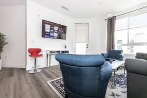 A modern living room with a flat-screen TV mounted on the wall displaying streaming services, a red bar stool, a blue armchair, and a grey sofa.