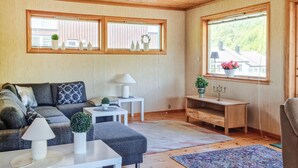 A cozy living room with a sectional sofa, a small side table, a lamp, and a candelabra. There are two windows with curtains, and a view of greenery outside. - Cozy home in Lyngdal with kitchen (Lyngdal)
