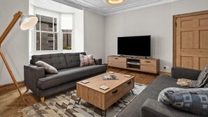 Een moderne woonkamer met een grijze bank, een houten salontafel, een televisie op een houten kast, en een groot raam met uitzicht op gebouwen.