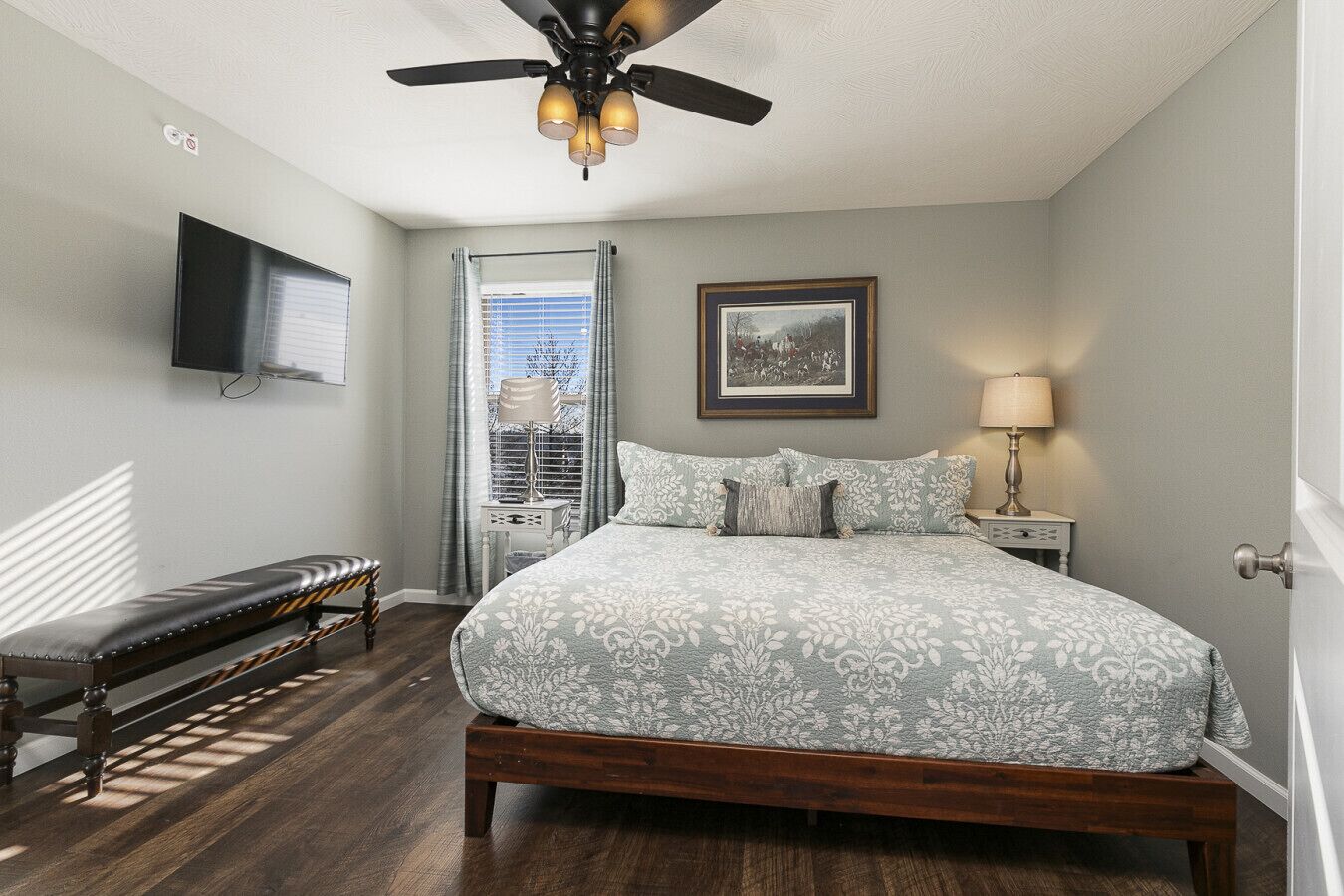 A bedroom with a bed, a bench, a ceiling fan, a TV, a window with curtains, and a framed picture on the wall.
