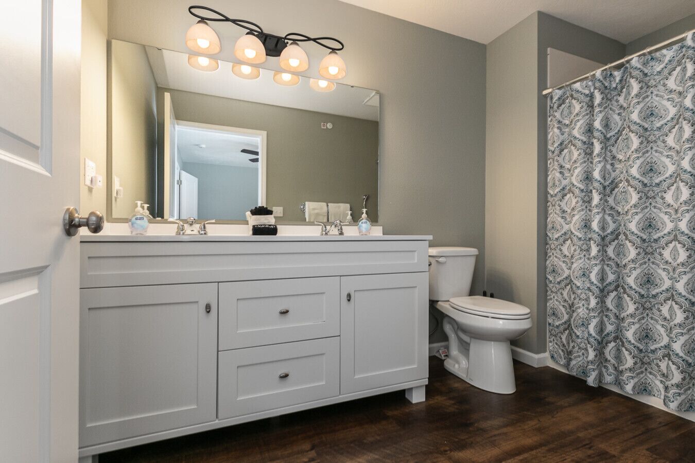 Une salle de bain avec un meuble-lavabo blanc, un miroir avec éclairage intégré, des toilettes et des rideaux de douche à motifs.