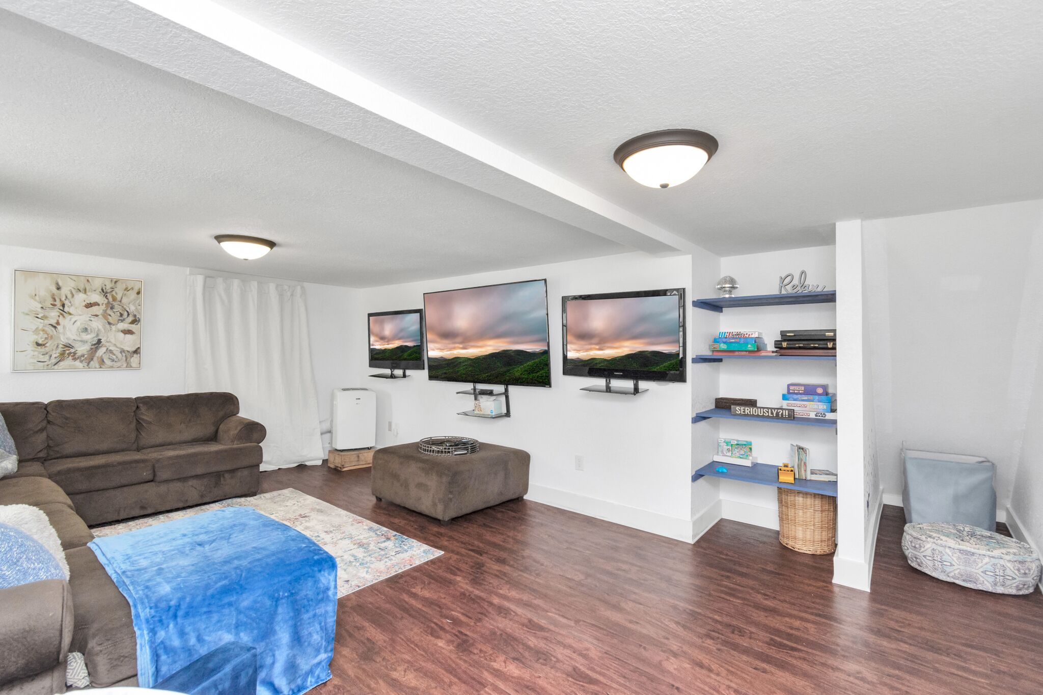 Una sala de estar moderna con un sofá marrón, un reposapiés gris y una manta azul. Hay tres televisores de pantalla plana montados en la pared, una estantería con libros y objetos decorativos, y un techo blanco con iluminación empotrada.