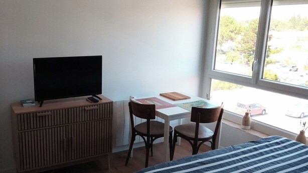 A room with a bed, a TV on a wooden cabinet, two chairs, a small table with books, and a window with a view of the street.