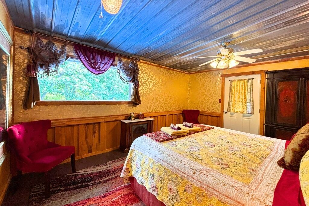 Une pièce comprenant un lit, un fauteuil rouge, une petite table, un ventilateur de plafond et une fenêtre avec des rideaux.
